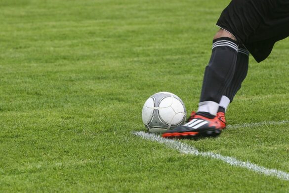Fußballspieler in schwarzen Stutzen und roten Schuhen tritt einen Ball auf einem grünen Spielfeld.