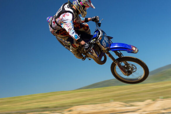 Motorradfahrer in buntem Outfit springt mit blauem Motorrad durch die Luft vor blauem Himmel.