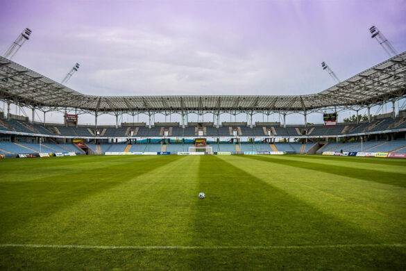Leeres Fußballstadion mit grüner Spielfläche, einem Ball in der Mitte und überdachten Tribünen im Hintergrund.