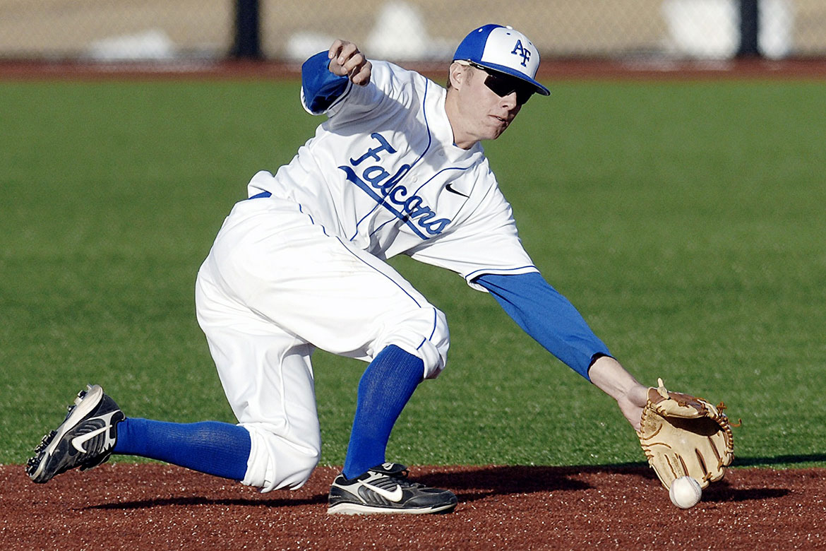 Baseballspieler in Aktion, bückt sich nach rechts und fängt einen rollenden Ball mit dem Handschuh auf einem Rasenplatz.